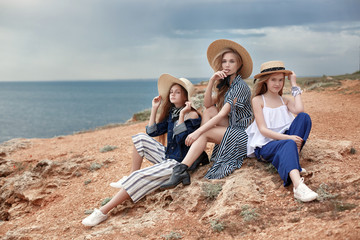 Beautiful, attractive and fun young girls having fun on the beach. Teenage sisters are resting together on the rocky shore of the blue ocean on a cloudy day against the blue sky and the coastline.