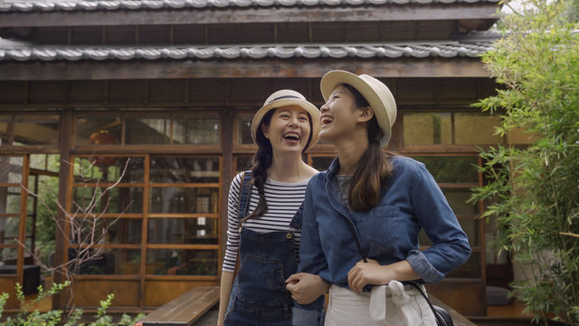summer holidays and vacation concept. smiling asian girls tourists walking close in japanese style garden laughing chatting having fun together. wooden house in background by teien on sunny day.