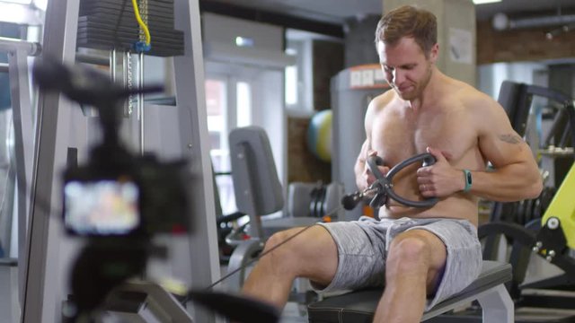 Rack focused shot of shirtless male coach with muscular body doing seated cable row, smiling and speaking on camera while recording online workout