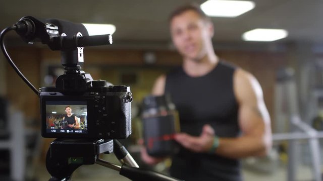 Rack focused shot of professional fitness coach looking at camera and telling about bodybuilding supplements while recording online commercial in gym