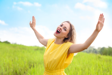 Happy young blonde woman in yellow dress is singing looking to camera on green meadow.