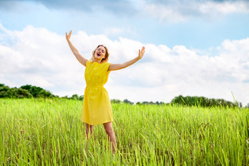 Naklejka na ściany i meble Happy young woman in yellow dress is singing with closed eyes and raising her hands on green meadow.
