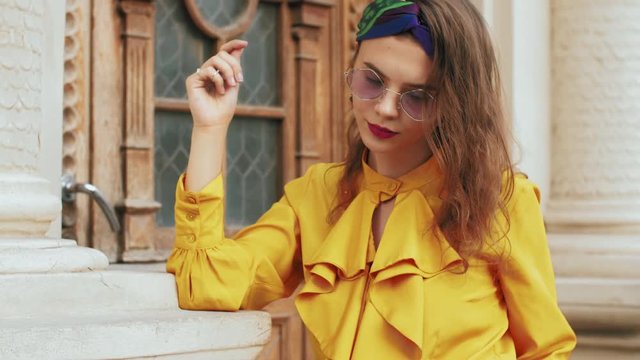 Street fashion portrait of young beautiful elegant lady wearing trendy purple color sunglasses, headband, stylish yellow blouse with jabot, posing in old european city