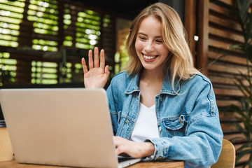 Happy young woman posing outdoors in cafe using laptop computer waving talking.