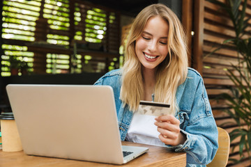Amazing excited happy young woman posing outdoors in cafe using laptop computer holding credit card.
