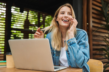 Excited happy young woman posing outdoors in cafe using laptop computer and talking by mobile phone.