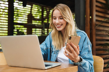 Excited happy young woman posing outdoors in cafe using laptop computer and mobile phone.