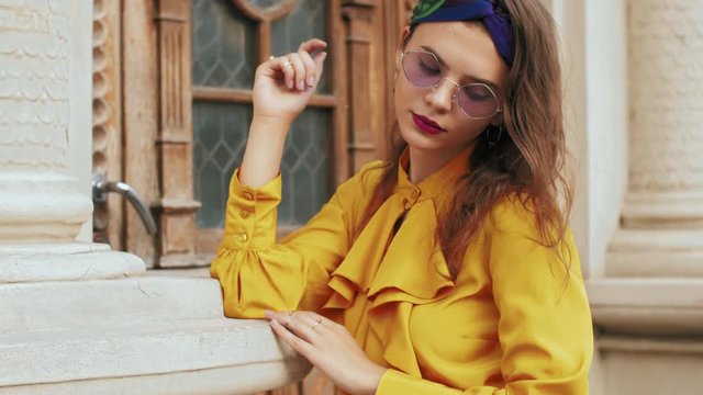 Street fashion portrait of young beautiful elegant woman wearing trendy purple color sunglasses, headband, stylish yellow blouse with jabot, posing in old european city