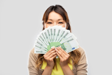 people, ethnicity and portrait concept - happy asian young woman holding hundreds of euro money banknotes over grey background