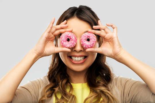 People, Fast Food And Fun Concept - Happy Asian Young Woman With Donuts Instead Of Eyes Over Grey Background