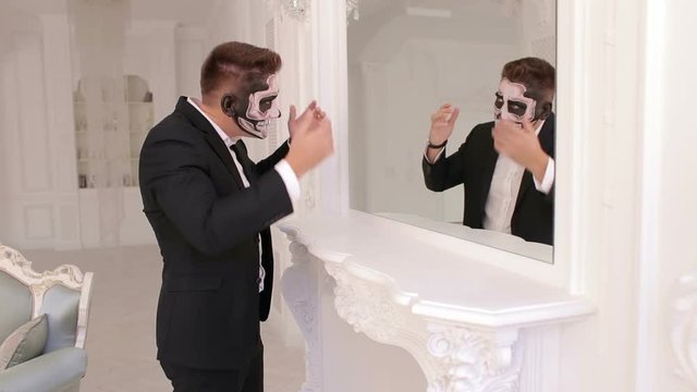 Creepy man businessman with makeup in the form of a skull stands in front of a mirror and straightens his suit. Halloween.