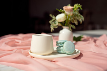Fototapeta na wymiar French blue macaroon and coffee cup standing on a wooden table with a pink tablecloth white vase with flowers roses and greens.