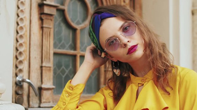 Street fashion portrait of young beautiful elegant lady wearing trendy purple color sunglasses, headband, stylish yellow blouse with jabot, posing in old european city
