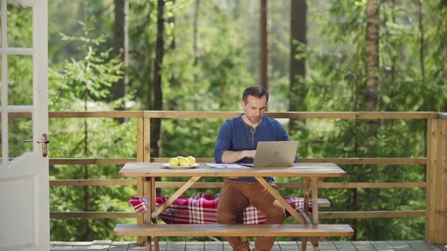 Tracking right shot of middle aged man typing on laptop computer and making notes on paper sitting at wooden table. Businessman working on country house porch surrounded by pine trees in summer