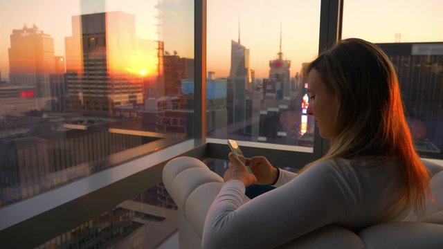 CLOSE UP: Caucasian girl looks at her smartphone from her office overlooking Times Square. Golden evening sunbeams shine on the towering skyscrapers surrounding girl texting from her hotel room.