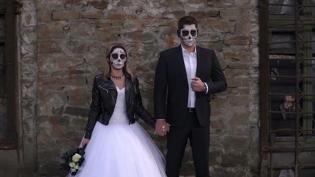 A horrible couple in a wedding dress and costume with makeup in the form of a skeleton stand against an old brick wall in an old abandoned house. The concept of Halloween. Halloween face art.