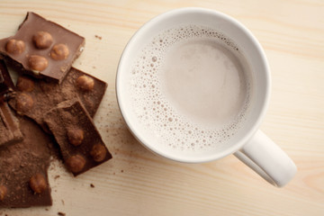 Cup with hot cocoa stands on wooden table near pieces of chocolate with nuts sprinkled with cocoa powder