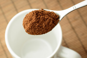 Spoon with ground coffee over the white empty cup against a brown bamboo background