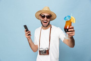 Excited young man tourist posing with camera using mobile phone drinking cocktail isolated over blue wall background.