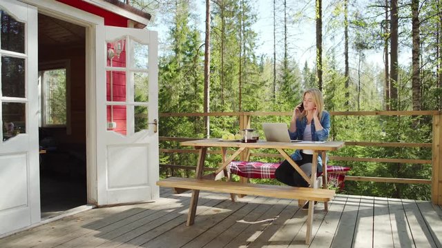 Tracking right shot of young woman talking on cell phone while working on laptop computer sitting on bench at wooden table on country house porch