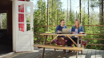 Tracking right shot of couple sitting at wooden table on country house balcony with open laptop and papers and planning family budget together over tea - Powered by Adobe