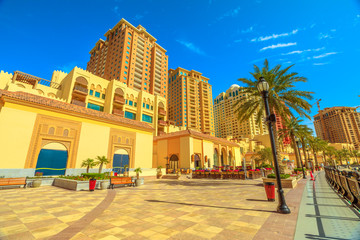 Fototapeta premium Benches and palm trees along marina walkway promenade in Porto Arabia at the Pearl-Qatar, Doha, with residential buildings towers on background. Persian Gulf in Middle East. Sunny day, blue sky.