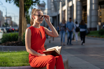 Fototapeta premium a young stylish european girl in a red suit and glasses sits on the parapet on the street and reads a book. Past her people pass, you can see the city life. There is a method for a small signature.