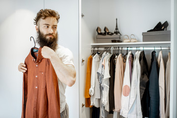 Bearded man trying shirt, choosing clothes to wear in the wardrobe at home
