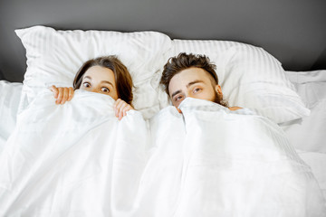 Young scared couple hiding under the bedsheets at home
