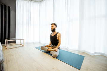 Fototapeta na wymiar Young bearded man practising yoga, feeling relaxed in the lotus position at the living room near the window at home