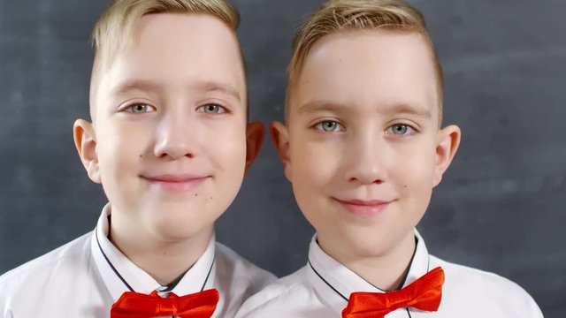 Close-up portrait shot of 10-year-old Caucasian identical twin brothers in smart white shirts and red bowties posing together against blackboard, looking at camera then smiling happily