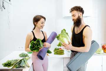 Portrait of a young couple in sportswear standing together with yoga mats and healthy fresh food on the kitchen at home
