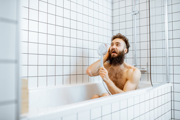 Joyful bearded man washing in the bathtub, having fun singing into the shower in the bathroom