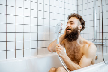 Joyful bearded man washing in the bathtub, having fun singing into the shower in the bathroom