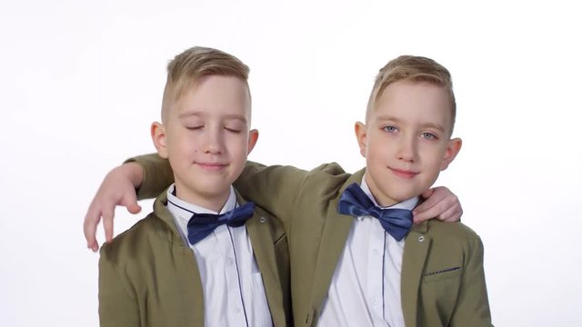Chest-up portrait shot of 9-year-old Caucasian twins with stylish haircuts, wearing smart blazers, white shirts and bowties, posing on white background, hugging each other’s shoulders and smiling