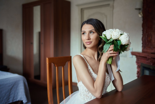 Beautiful bride with the peony bouquet is sitting in the hotel&rsquo;s room near the mirror.