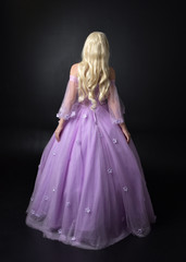 full length portrait of a blonde girl wearing a fantasy fairy inspired costume,  long purple ball gown with fairy wings,   standing pose  on a dark studio background.