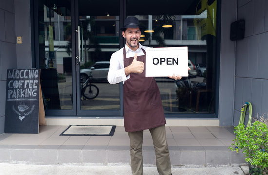 Portrait of a happy waitress standing at coffee shop entrance and holding open sign in front coffee shop. Startup Small Business owner concept. SME Business Concept.