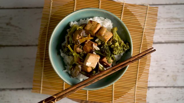 Turning shot of bowl of stir fried tofu and broccoli with rice on sushi mat and plain painted wood background.
