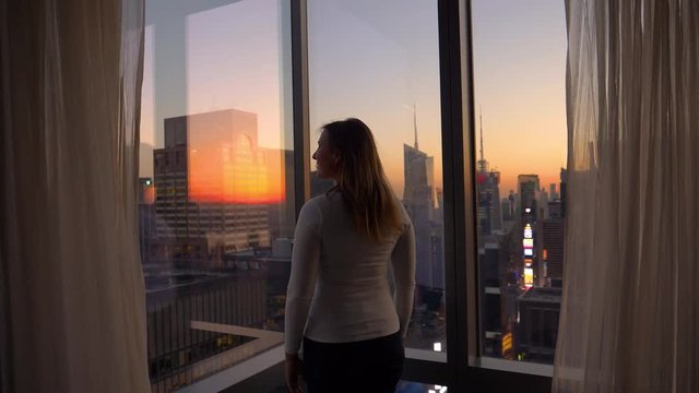 CLOSE UP: Cheerful Caucasian woman opens the curtains and looks at the metropolitan city at sunset. Happy female traveler draws the curtains to observe the breathtaking Times Square on a sunny evening