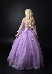 Fototapeta premium full length portrait of a blonde girl wearing a fantasy fairy inspired costume, long purple ball gown with fairy wings, standing pose on a dark studio background.