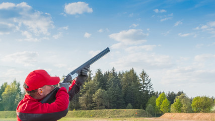 trap or skeet shooting, man in red clothes  shoots from a shotgun at clay pigeon,  a background of forest and sky, Images with Copy Space - obrazy, fototapety, plakaty