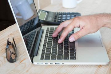 man's hand on the laptop keyboard, there is a calculator and glasses on the table