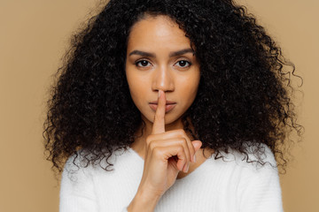 Image of serious confident dark skinned woman makes shush gesture, keeps fore finger over lips,...