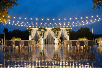 Night wedding ceremony with arch, orchid flowers, chairs and bulb lights in forest outdoors, copy space. Wedding decorations