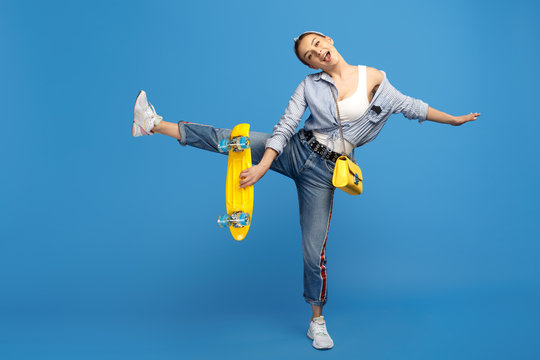 Full length photo of happy young woman with yellow penny or skateboard dancing over blue background.