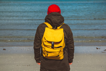 Traveler man with a yellow backpack wearing a red hat standing on a sandy beach on the background of the sea. Shoot from the back
