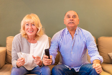 Multiracial senior couple fans cheering for soccer (football), watching TV show on sofa