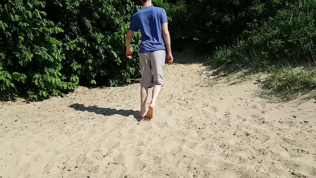 The man can hardly walk on the sand with his bare feet. The view from the back. Lameness. Walk-treatment of a person with cerebral palsy. Difficulties of people with disabilities
