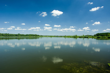 Calm lake, trees on the horizon and white clouds on a blue sky reflecting in the water